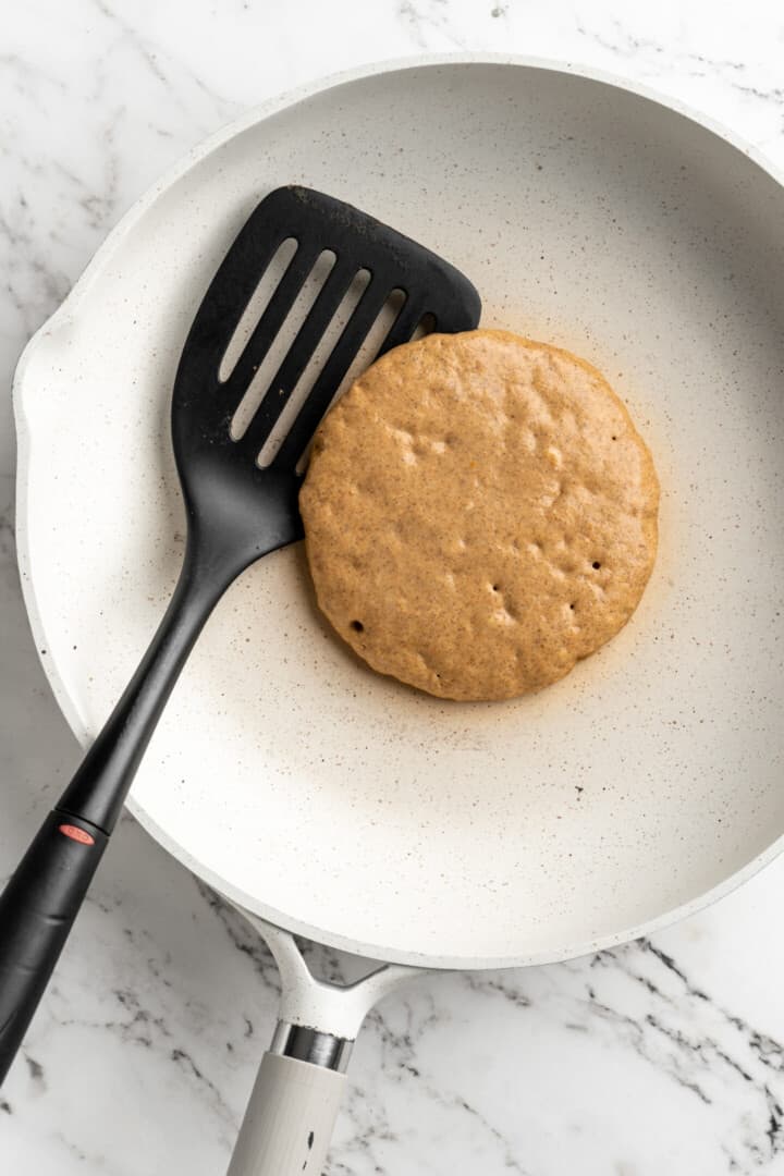 Overhead view of an uncooked pumpkin pancake in a pan with a spatula.