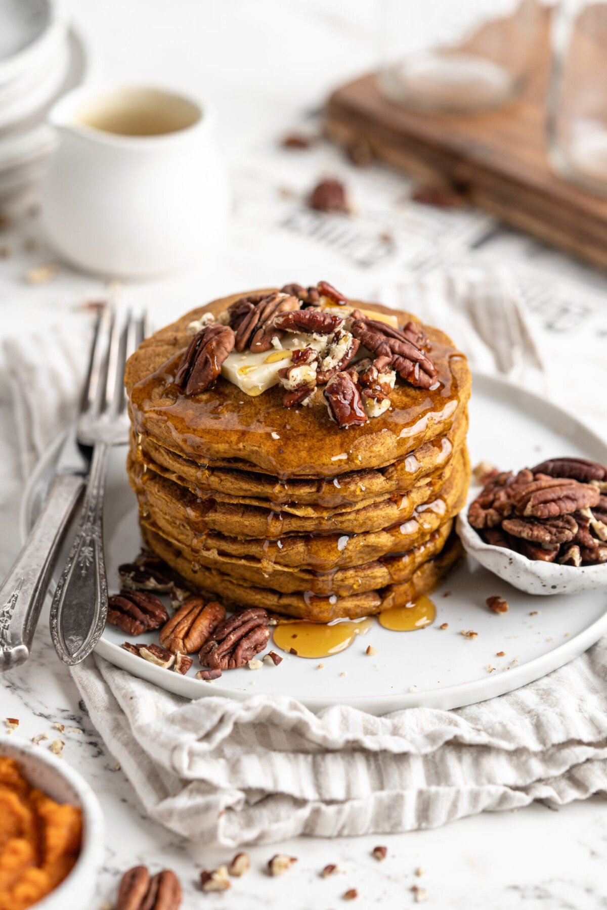 Overhead view of a stack of pumpkin pancakes on a plate, topped with nuts, next to a bowl of nuts and a fork.