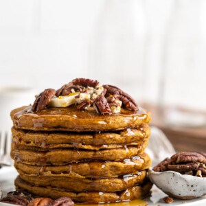 A stack of pumpkin pancakes on a plate, topped with nuts, next to a bowl of nuts.