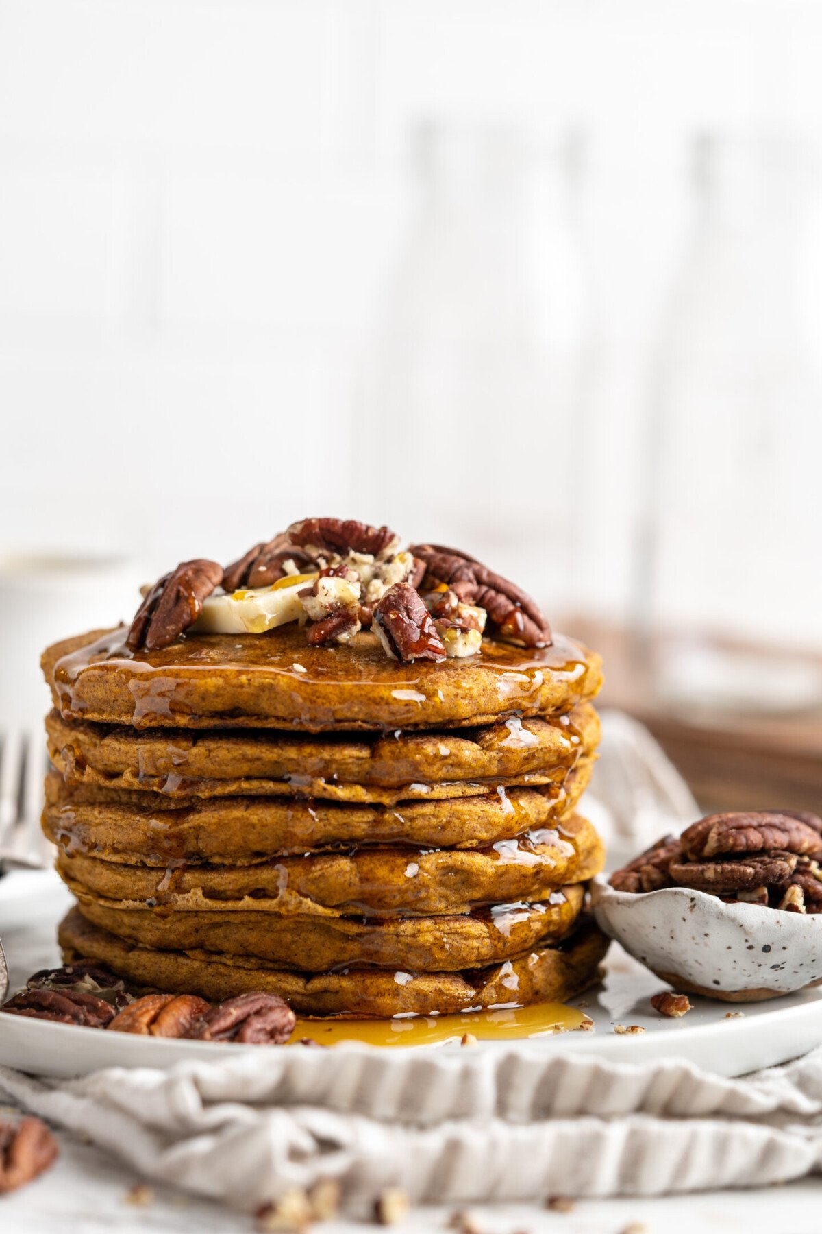 A stack of pumpkin pancakes on a plate, topped with nuts, next to a bowl of nuts.