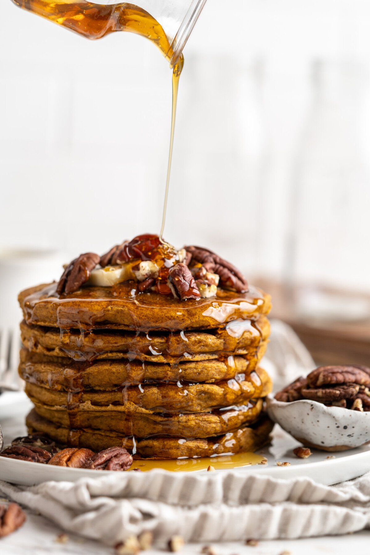 A stack of pumpkin pancakes on a plate with nuts, with syrup being poured over the top.