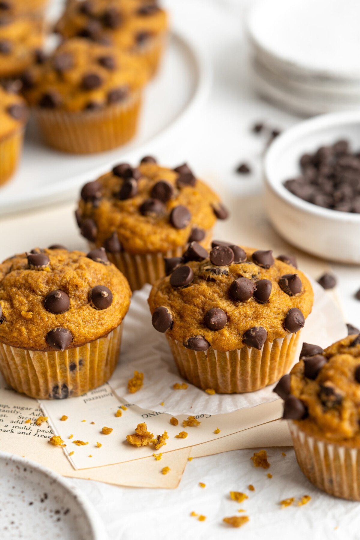 Four pumpkin chocolate chip muffins with a plate of muffins in the background, and a bowl of chocolate chips.