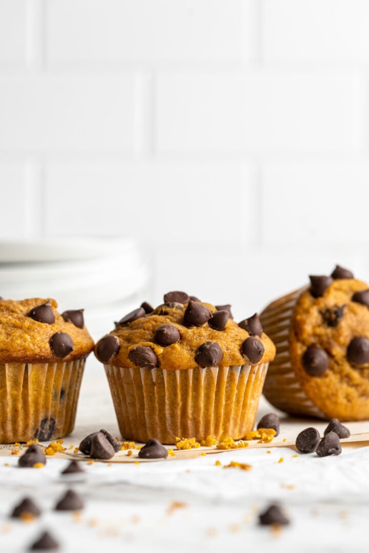 Three pumpkin chocolate chip muffins in a row, surrounded by chocolate chips.
