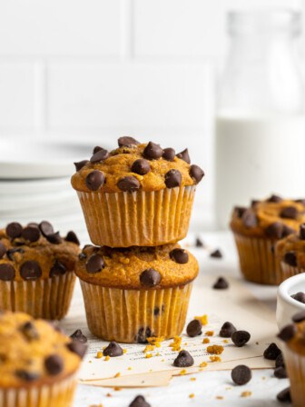 Two chocolate chip pumpkin muffins stacked on top of each other, surrounded by chocolate chips, with a glass of milk in the background.