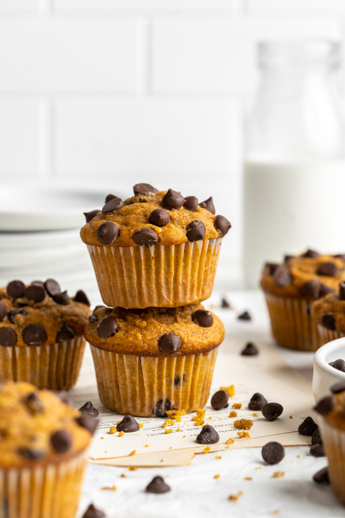 Two chocolate chip pumpkin muffins stacked on top of each other, surrounded by chocolate chips, with a glass of milk in the background.