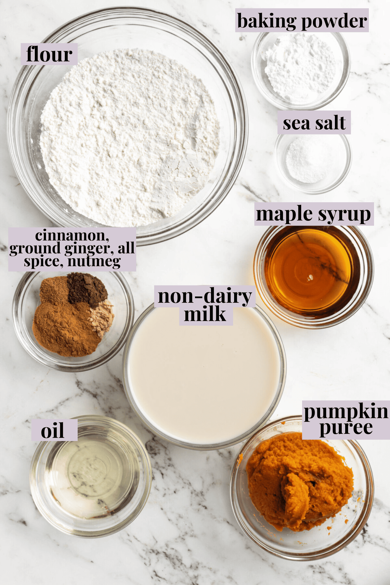 Overhead view of the ingredients needed for gluten free pumpkin pancakes: a bowl of oat flour, a glass of almond milk, a bowl of salt, a bowl of baking powder, a bowl of cinnamon, a bowl of maple syrup, a bowl of pumpkin puree, and a bowl of oil.