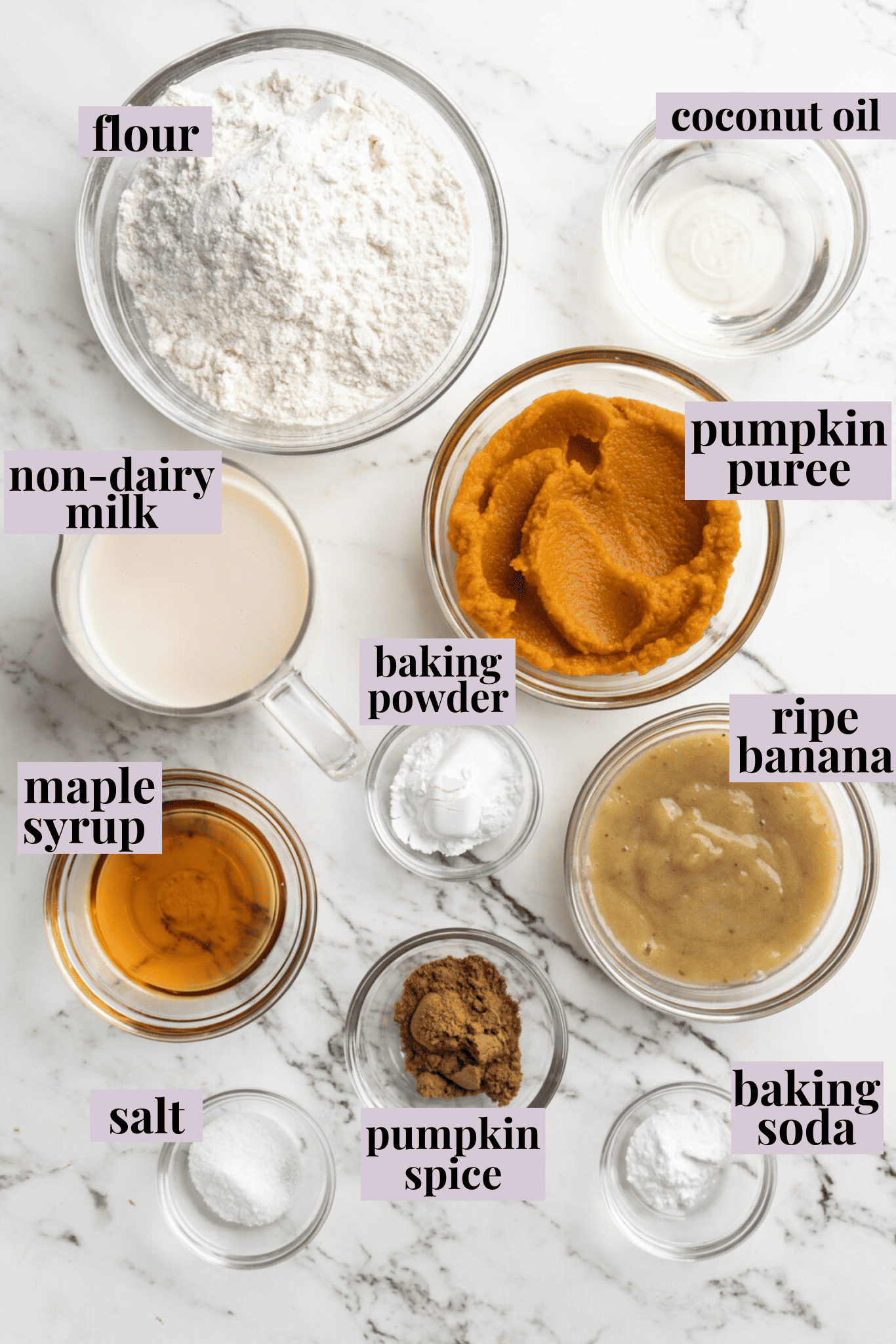 Overhead view of the ingredients needed for pumpkin waffles: a bowl of flour, a bowl of pumpkin puree, a pyrex of non-dairy milk, a glass of agave, a bowl of mashed banana, a glass of coconut oil, a bowl of pumpkin spice, a bowl of salt, a bowl of baking powder, and a bowl of baking soda