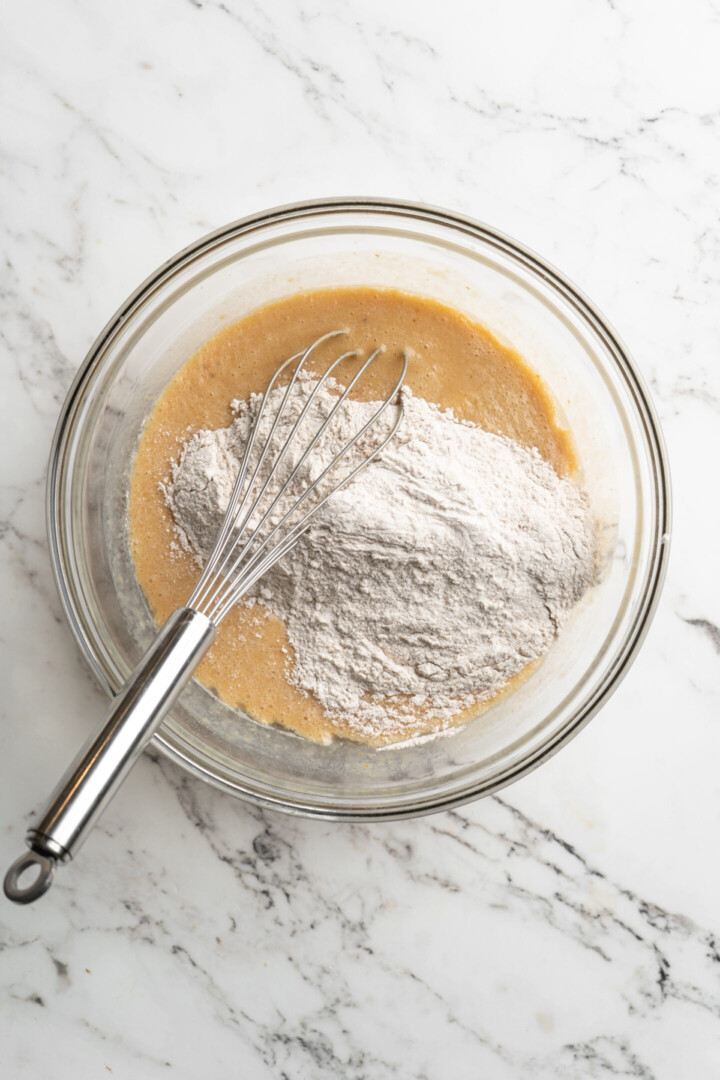 A mixing bowl with a pile of flower on top of wet ingredients, with a whisk