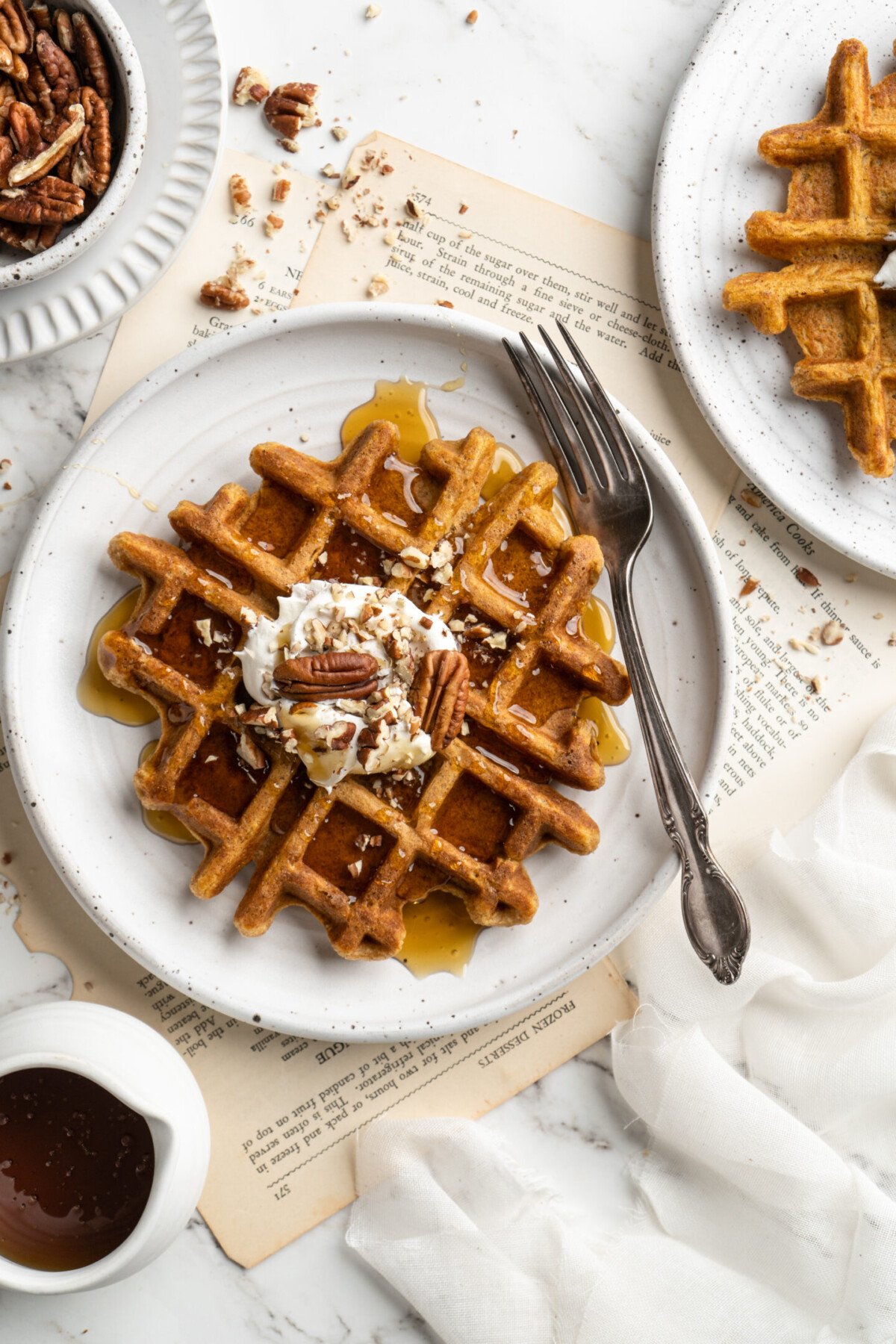 Overhead view of a plate with a fork, a pumpkin waffle, a dollop of whipped coconut cream, and candied pecans, next to a plate with anotherwaffle