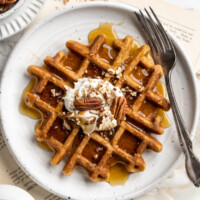 Overhead view of a pumpkin waffle on a plate with a fork, topped with coconut cream and pecans