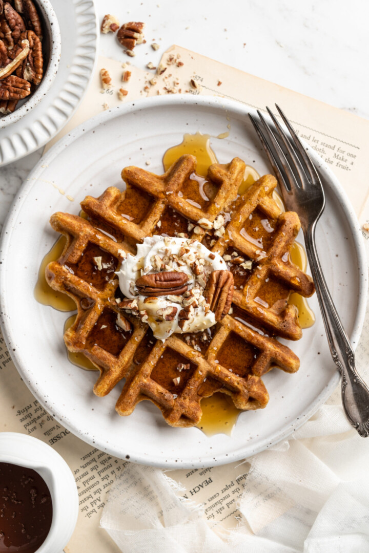 Overhead view of a pumpkin waffle on a plate with a fork, topped with coconut cream and pecans