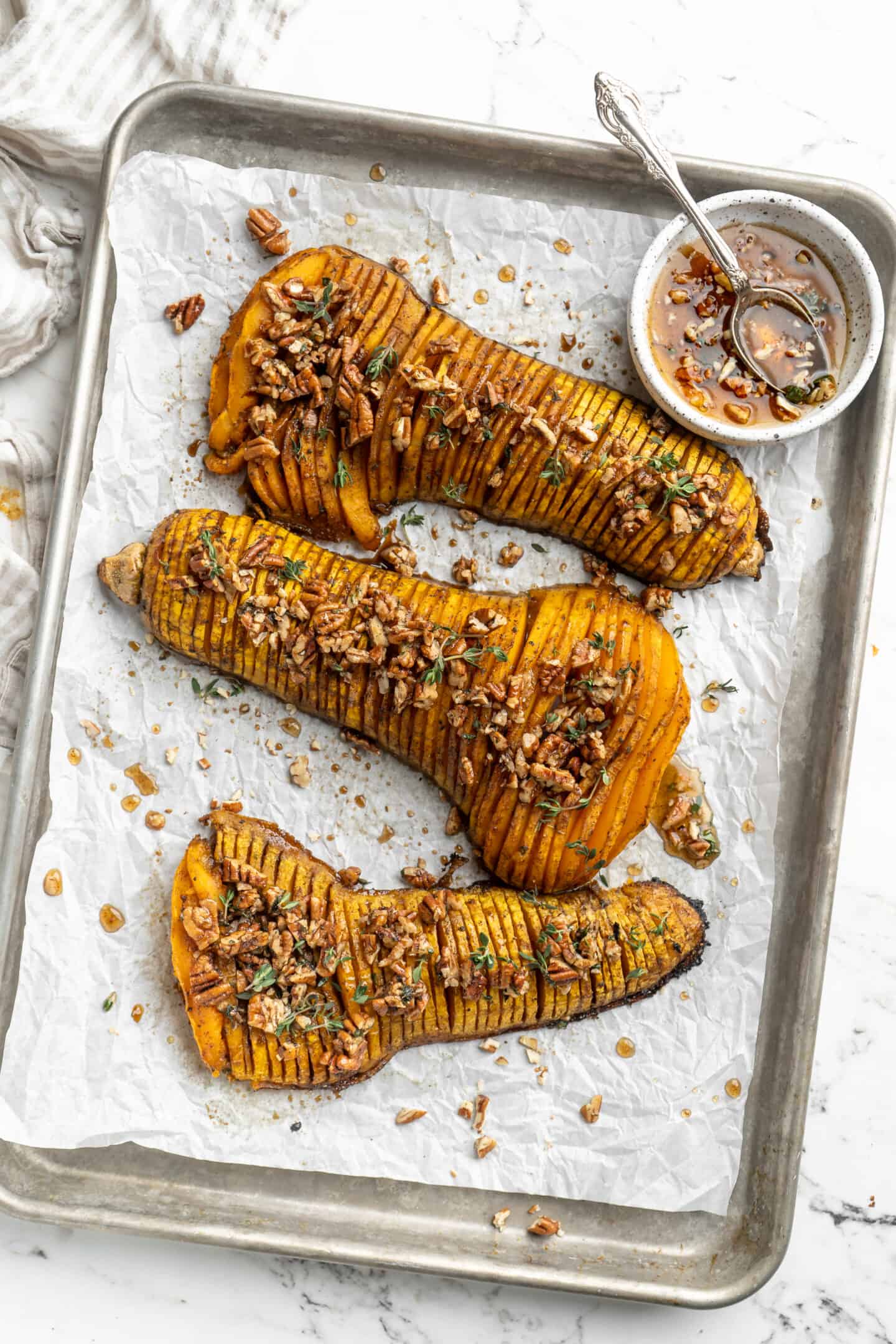 Overhead view of hasselback butternut squash on parchment-lined baking sheet with maple pecan glaze in small bowl