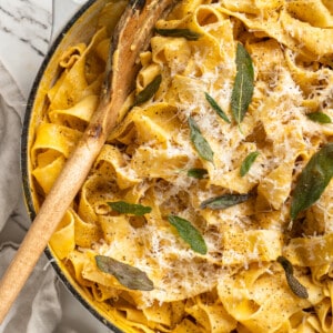 Overhead view of pumpkin alfredo pasta in skillet with wooden spoon and crispy sage leaf garnish