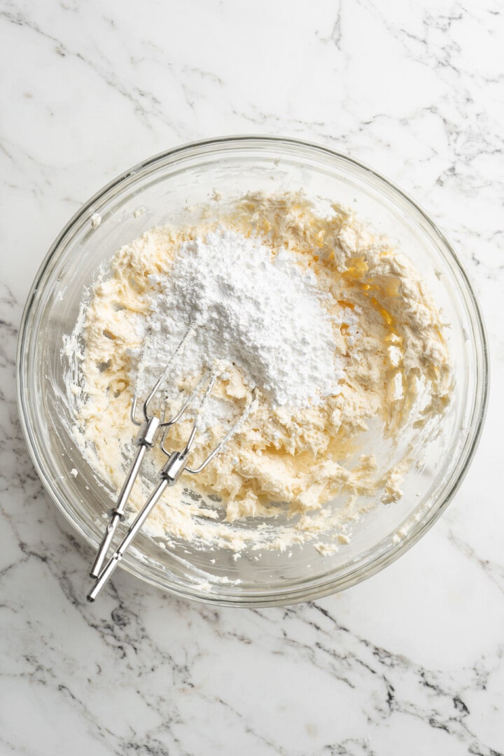 Overhead view of powdered sugar added to bowl of creamed butter and cream cheese
