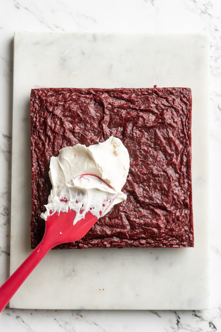 Spatula spreading cream cheese frosting onto red velvet brownies