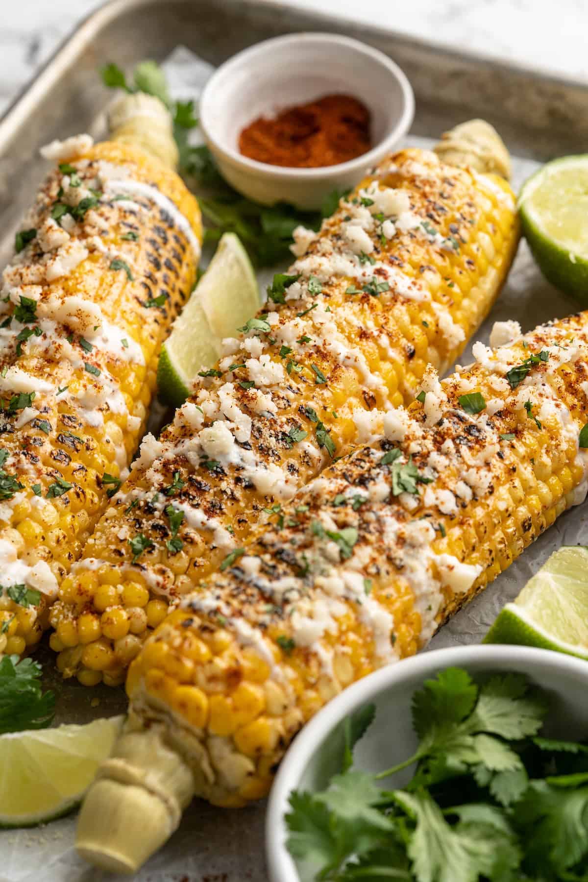 Elotes on a sheet pan with bowls of chili powder, cilantro, and lime wedges