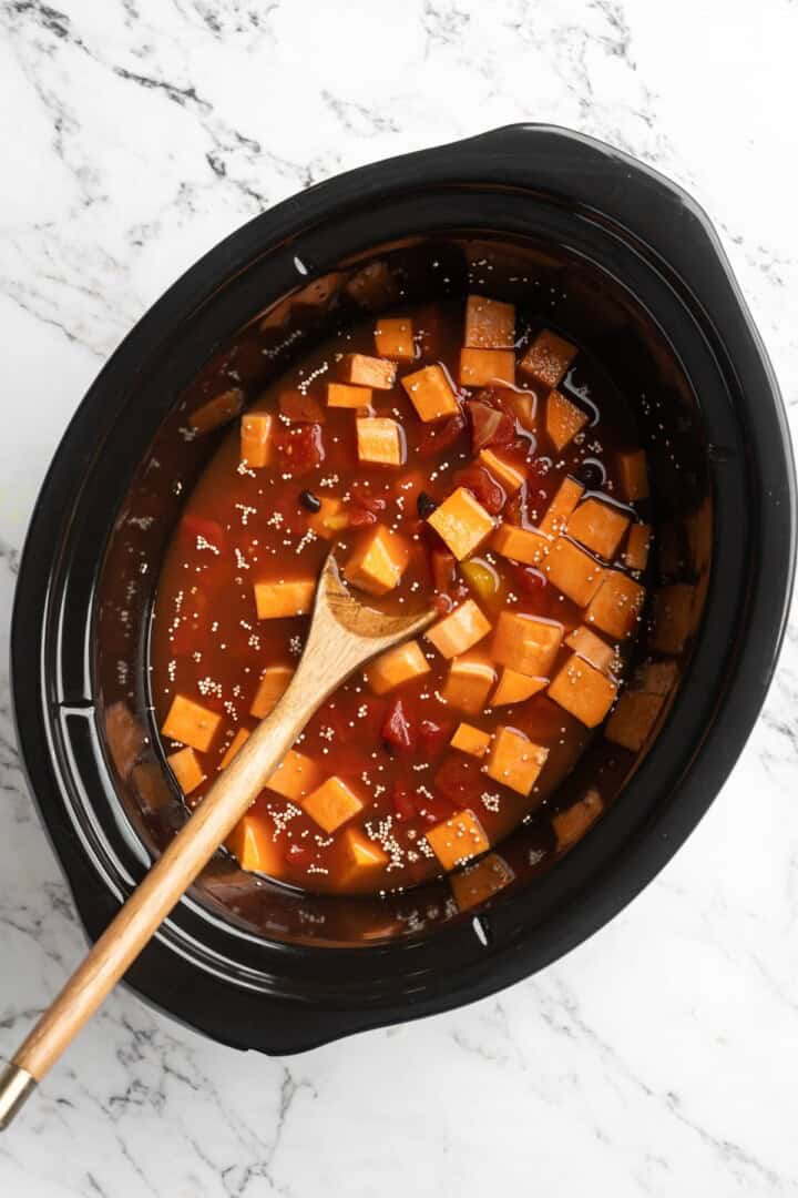 Overhead view of sweet potatoes, black beans, tomatoes, and broth in slow cooker insert with wooden spoon