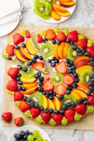 Overhead view of whole fruit pizza on parchment with piece cut and pulled away