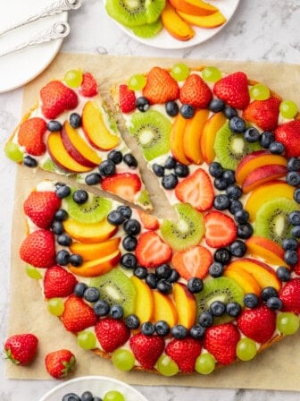 Overhead view of whole fruit pizza on parchment with piece cut and pulled away