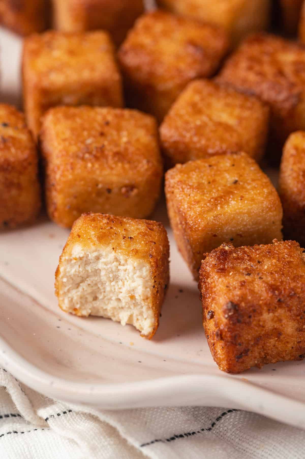 Closeup of fried tofu on plate, with one cube bitten to show soft interior