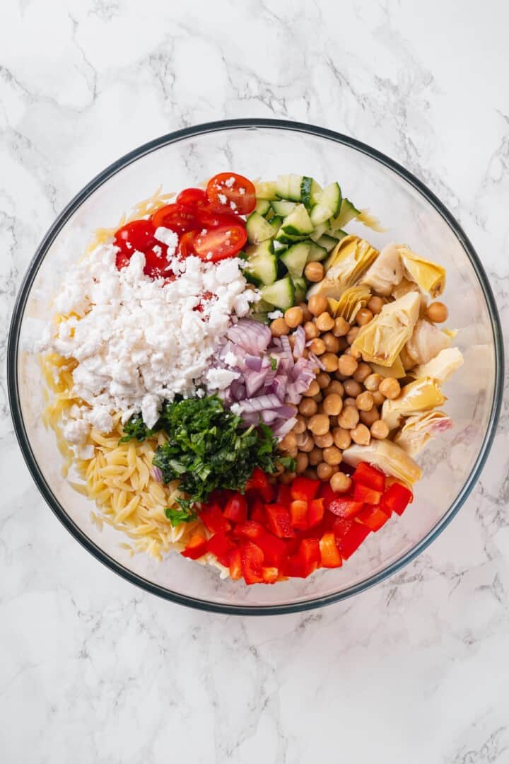 Overhead view of ingredients for orzo pasta salad in bowl before tossing