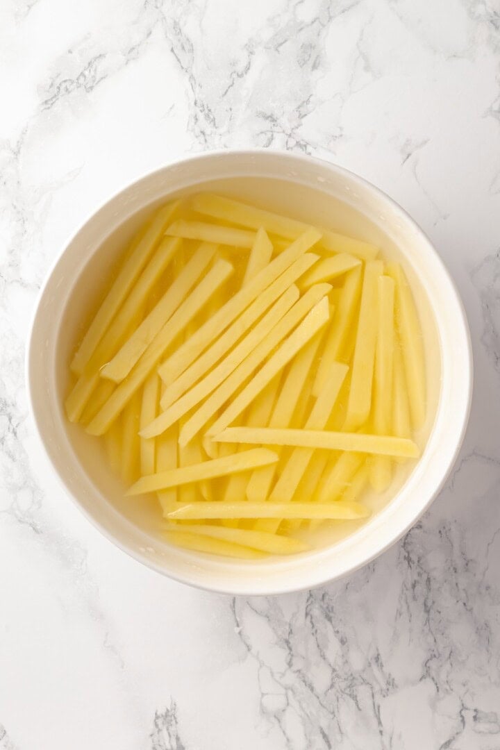 Overhead view of raw potatoes cut into fries and soaking in bowl of water