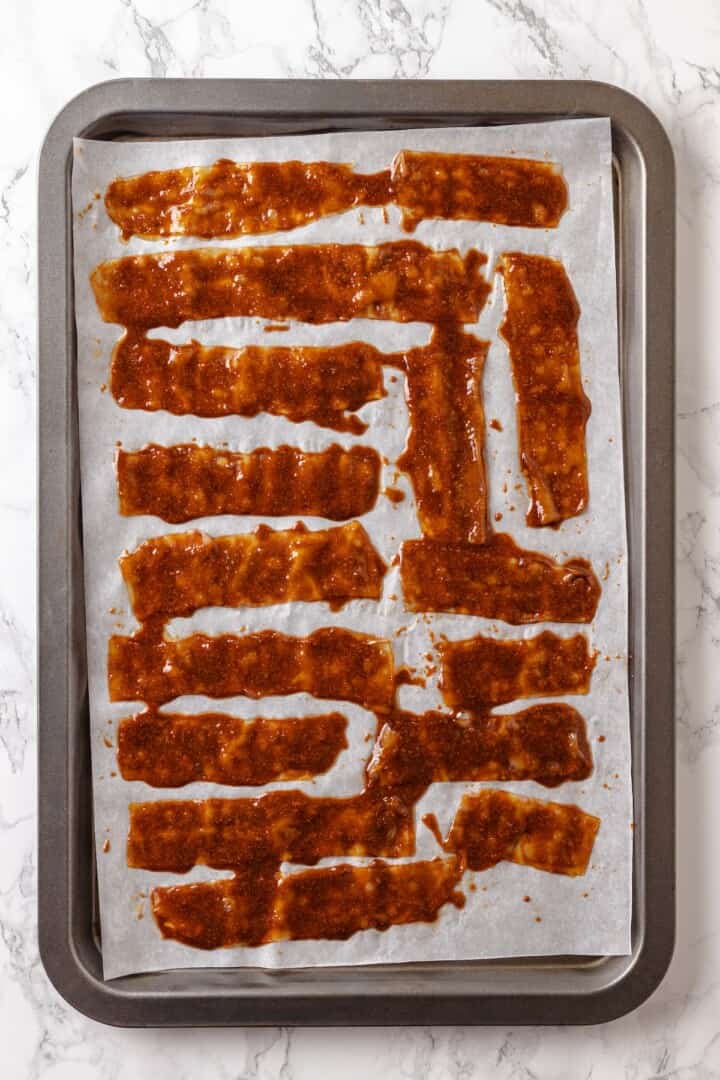 Rice paper is the secret for ultra crispy vegan bacon! This recipe is smoky, sweet, and savoury, and it's incredibly easy to make too. Overhead view of vegan rice paper bacon on parchment-lined baking sheet halfway through baking
