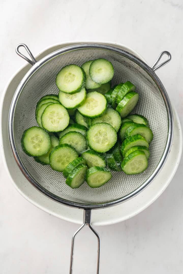 Overhead view of salted cucumber slices in colander set over bowl
