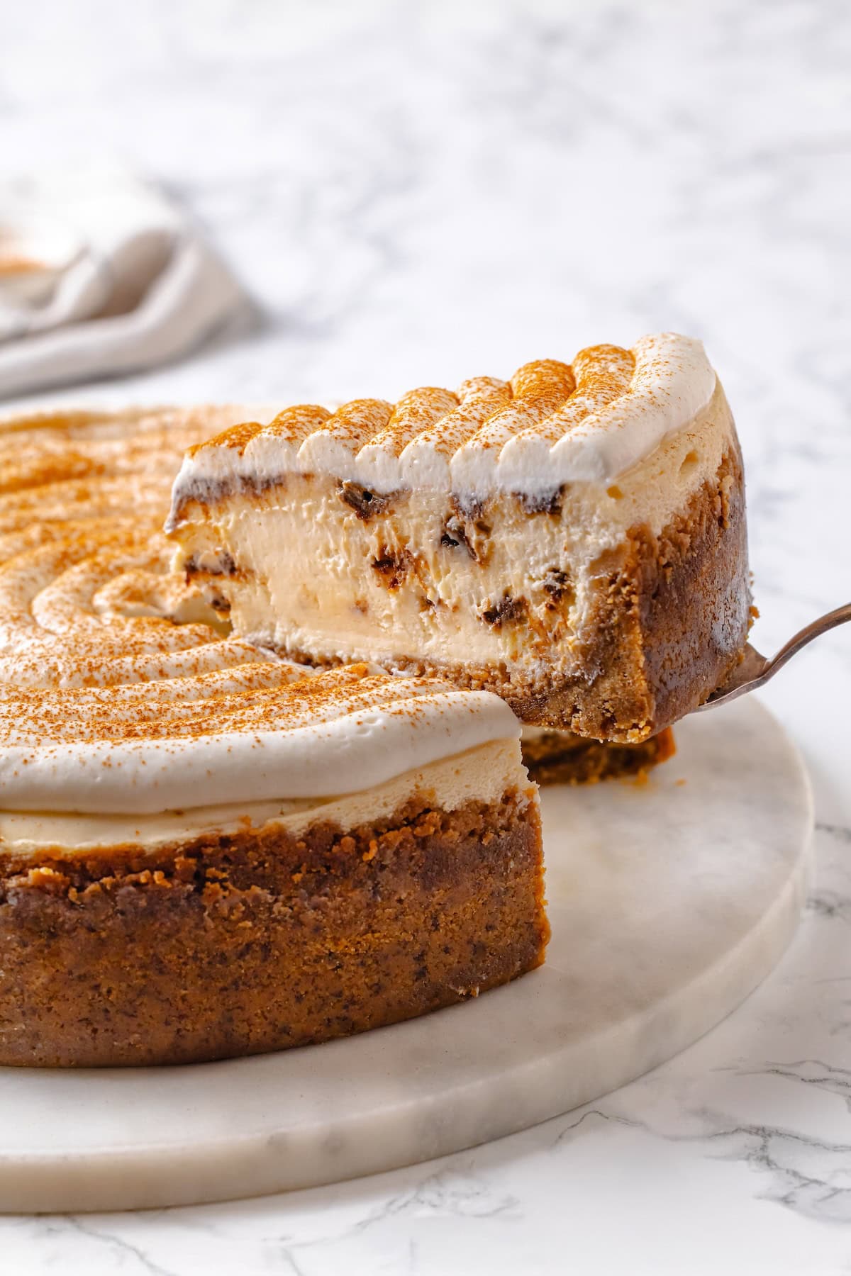 With a brown sugar cinnamon swirl and cream cheese frosting, this cinnamon roll cheesecake recipe is cozy and decadent at the same time! Lifting slice of cinnamon roll cheesecake from marble platter