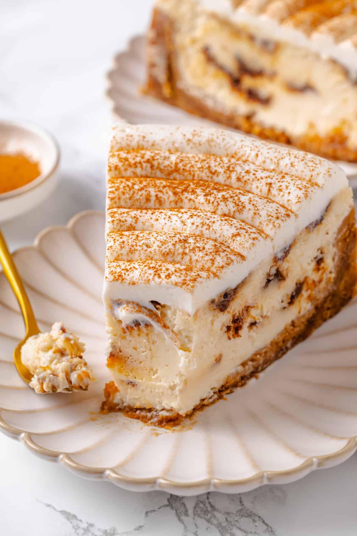With a brown sugar cinnamon swirl and cream cheese frosting, this cinnamon roll cheesecake recipe is cozy and decadent at the same time! Slice of cinnamon roll cheesecake on plate with tip on fork