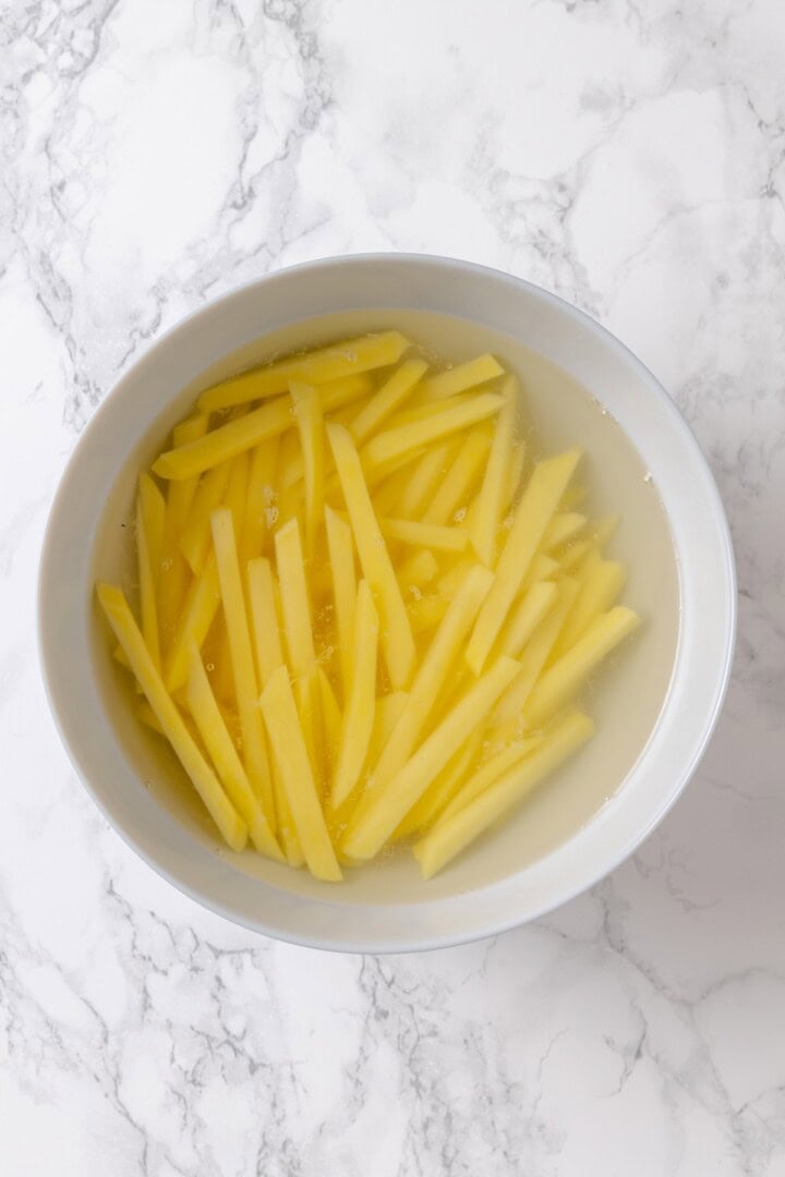 Whether you bake them or deep fry them, these crispy Garlic Fries are guaranteed to be a hit! They're cheesy, easy and packed with flavour. Overhead view of fries soaking in bowl of cold water