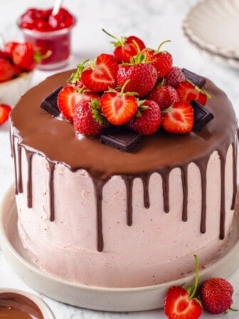 Chocolate strawberry cake on cake plate with ganache, berries, and chocolate pieces on top