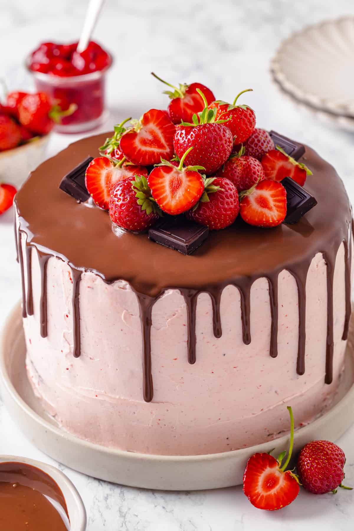 Fluffy chocolate cake, a jammy strawberry filling, strawberry buttercream and chocolate ganache make this Chocolate Strawberry Cake the best! Chocolate strawberry cake on cake plate with ganache, berries, and chocolate pieces on top