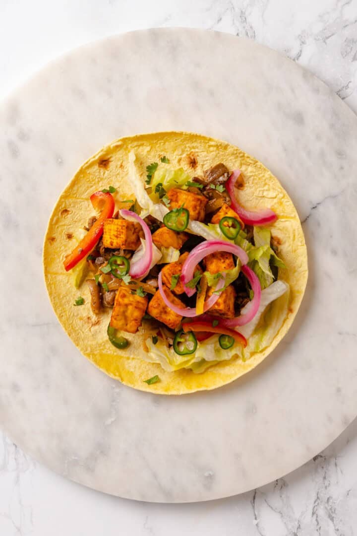 These crispy Tofu Tacos are topped with meaty fajita mushrooms and veggies for a vegan taco recipe that’s satisfying and delicious! Overhead view of taco with tofu, fajitas, and pickled onions