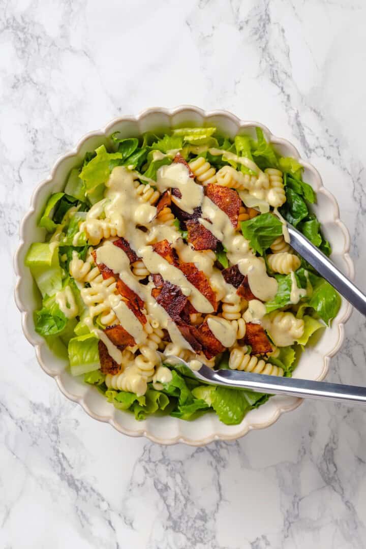 Tongs tossing caesar pasta salad in mixing bowl.