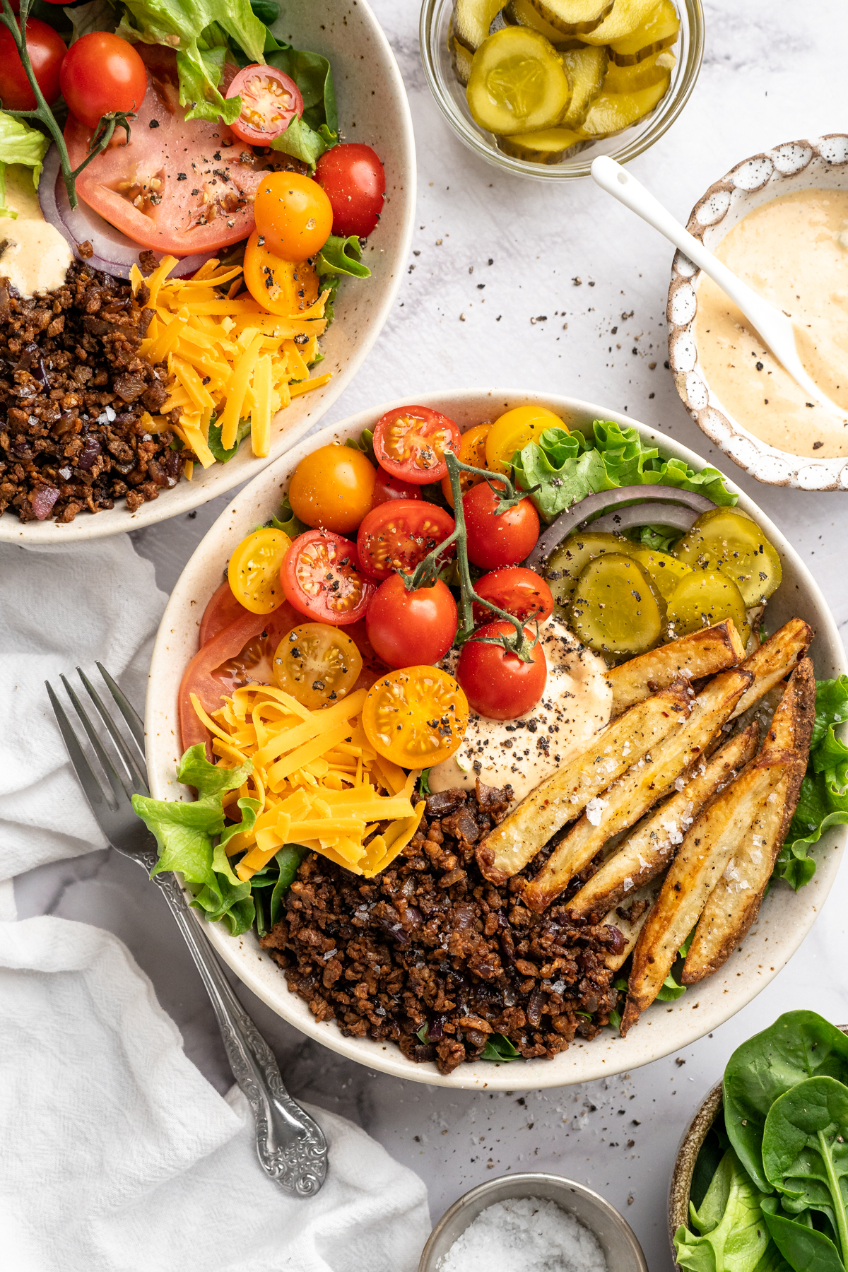 Two vegan cheeseburger bowls with tomatoes, fries, pickles, and other toppings.