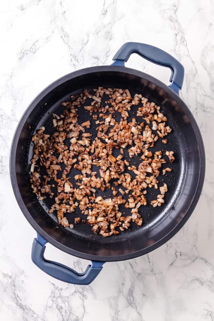 Warm spices mingle with vegetables, golden raisins, olives and chickpeas to make this Moroccan Cauliflower Tagine a delicious vegan dinner! Onions, garlic, and spices cooking in pan.