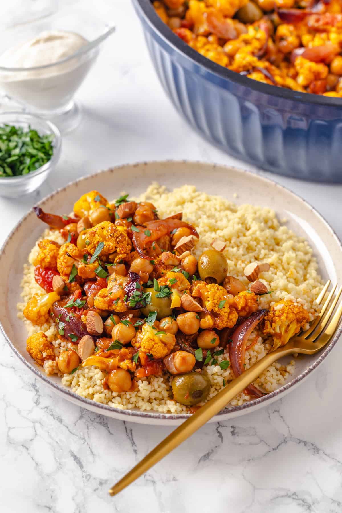 Warm spices mingle with vegetables, golden raisins, olives and chickpeas to make this Moroccan Cauliflower Tagine a delicious vegan dinner! Bowl of Moroccan cauliflower tagine with couscous and gold fork.