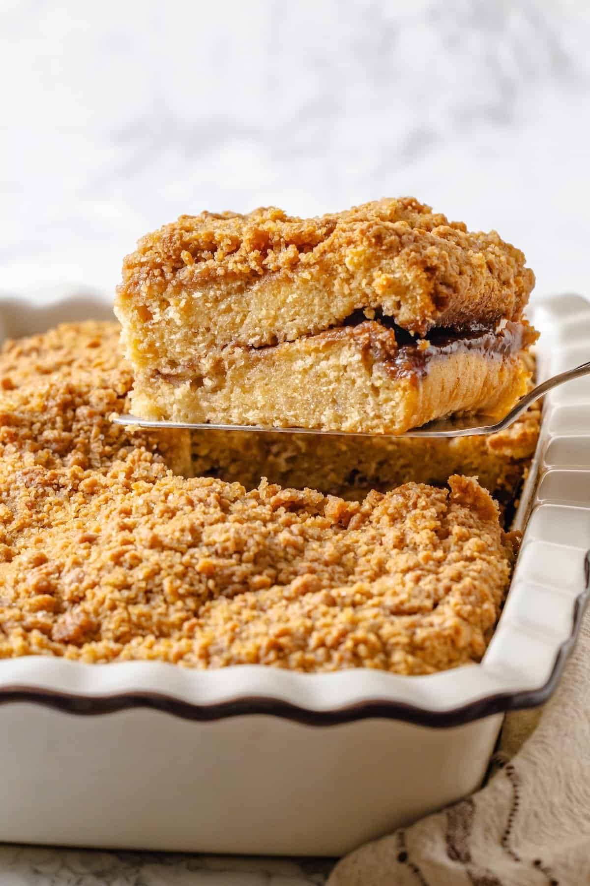 This vegan coffee cake recipe is a bakery-style treat! Moist and tender, with a cinnamon swirl and irresistible crumb topping. Delicious! Spatula lifting slice of vegan coffee cake from baking dish.