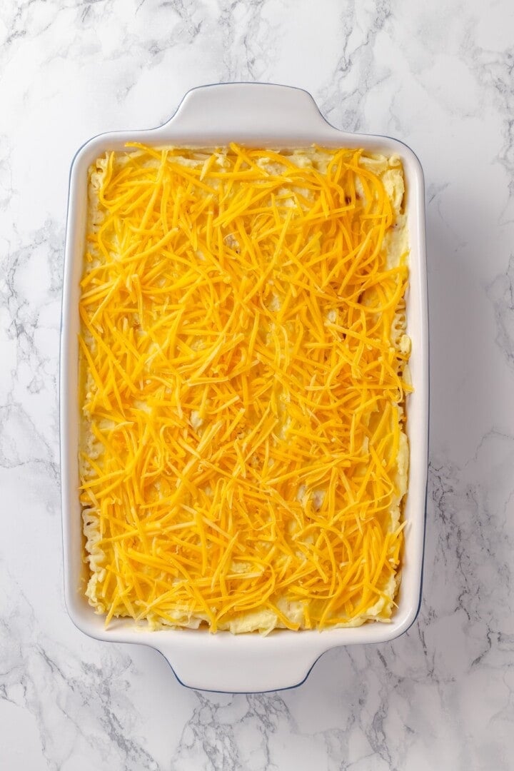 This vegan pierogi casserole layers a cheesy potato filling with lasagna noodles for pierogi vibes without the work of making dumplings! Vegan cheese added to pierogi casserole in baking dish.