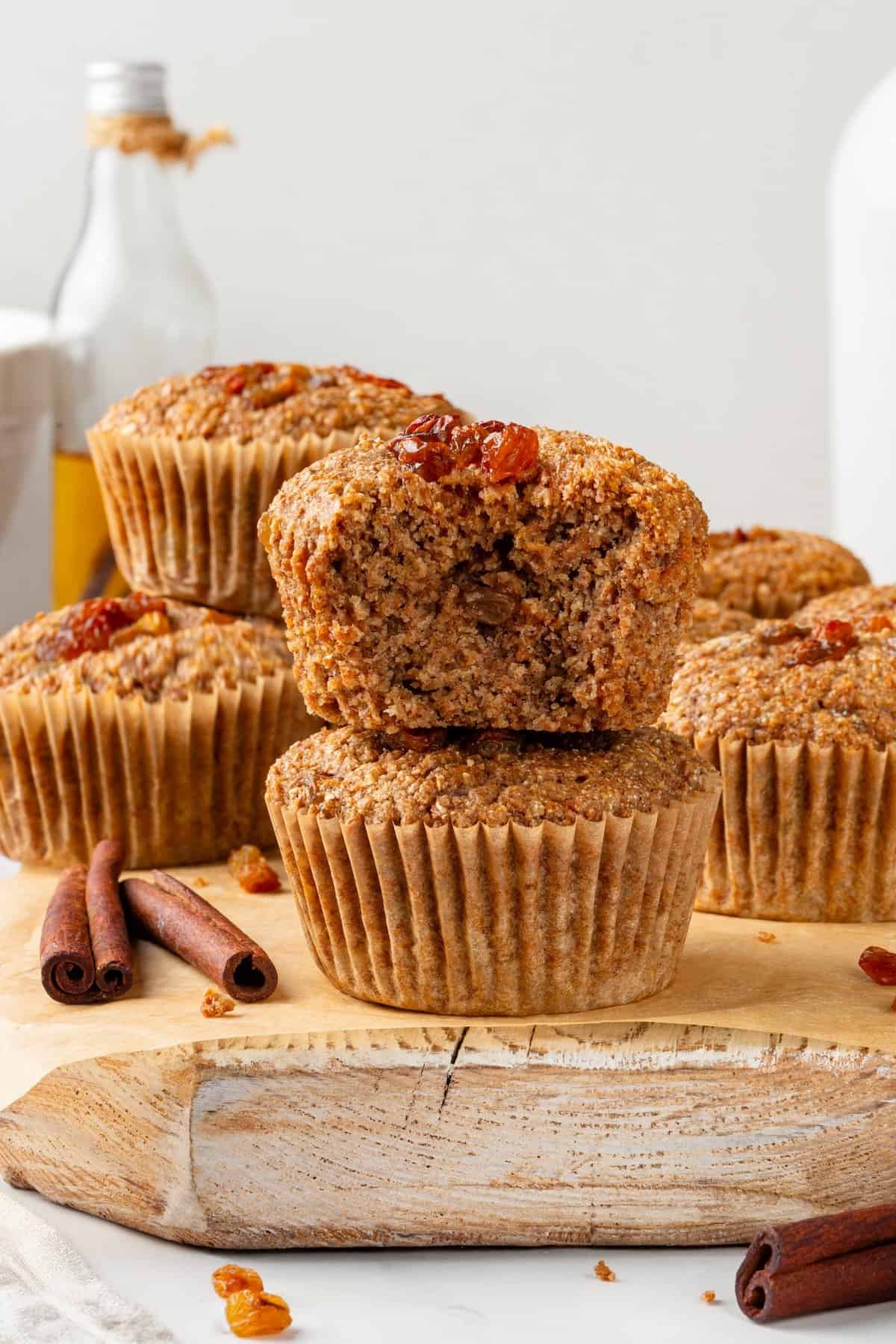 This moist vegan bran muffin recipe is made with whole wheat flour, raisins, and spiced with cinnamon for an easy, wholesome breakfast! Stack of 2 bran muffins with top bitten to show tender crumb and additional muffins in background.