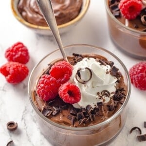 Spoon in bowl of silken tofu chocolate mousse with vegan whipped cream, raspberries, and chocolate shavings.