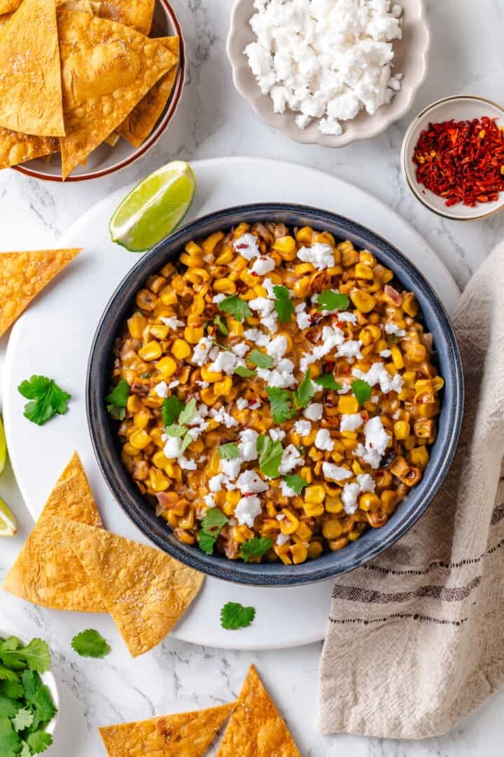 This vegan Mexican Street Corn Dip is creamy, smoky, and a little bit spicy (just like elotes!) for an easy appetizer everyone will love! Overhead view of Mexican street corn dip in bowl with bowls of tortilla chips, vegan cotija, and chili flakes.