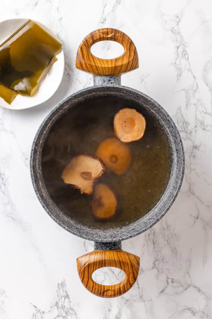 This cozy Miso Tofu Soup recipe is made with wakame, ginger, carrots and homemade dashi for a light, nourishing and flavorful vegan soup. Shiitake mushrooms soaking in pot of broth set next to plate of kombu.