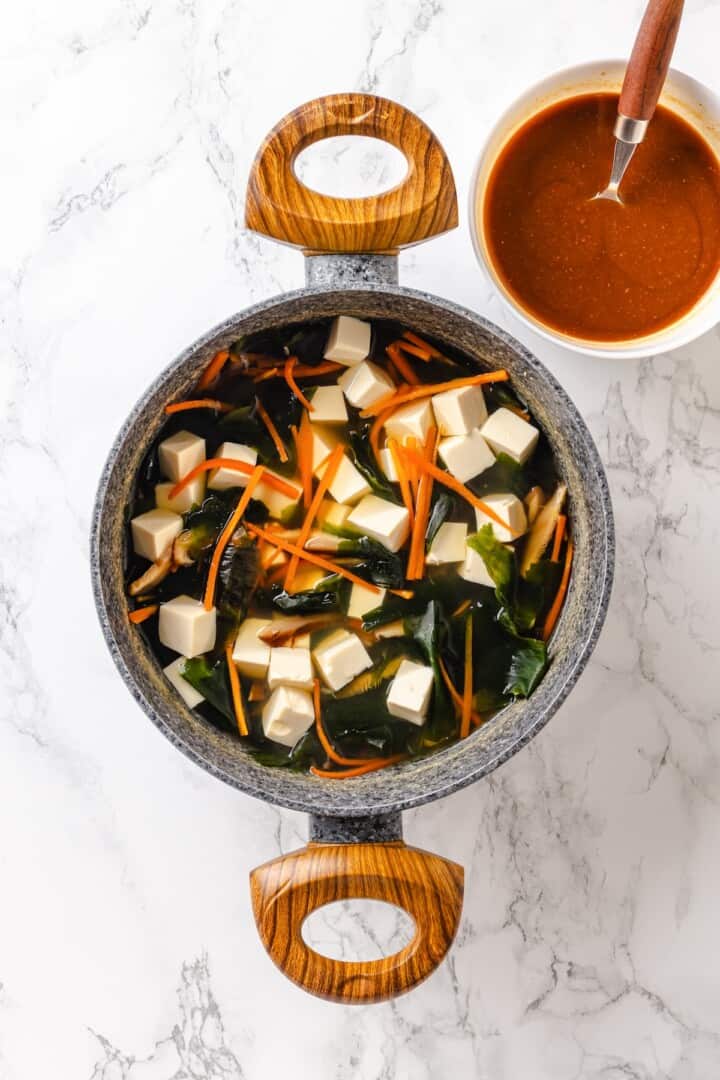 This cozy Miso Tofu Soup recipe is made with wakame, ginger, carrots and homemade dashi for a light, nourishing and flavorful vegan soup. Vegan miso tofu soup set next to bowl of miso and broth mixture.