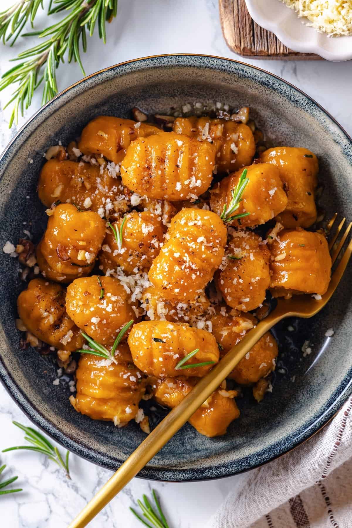 This soft, pillowy homemade sweet potato gnocchi is tossed with rosemary garlic butter and walnuts for a restaurant-style vegan dinner! Fork in bowl of sweet potato gnocchi with vegan Parmesan and rosemary.
