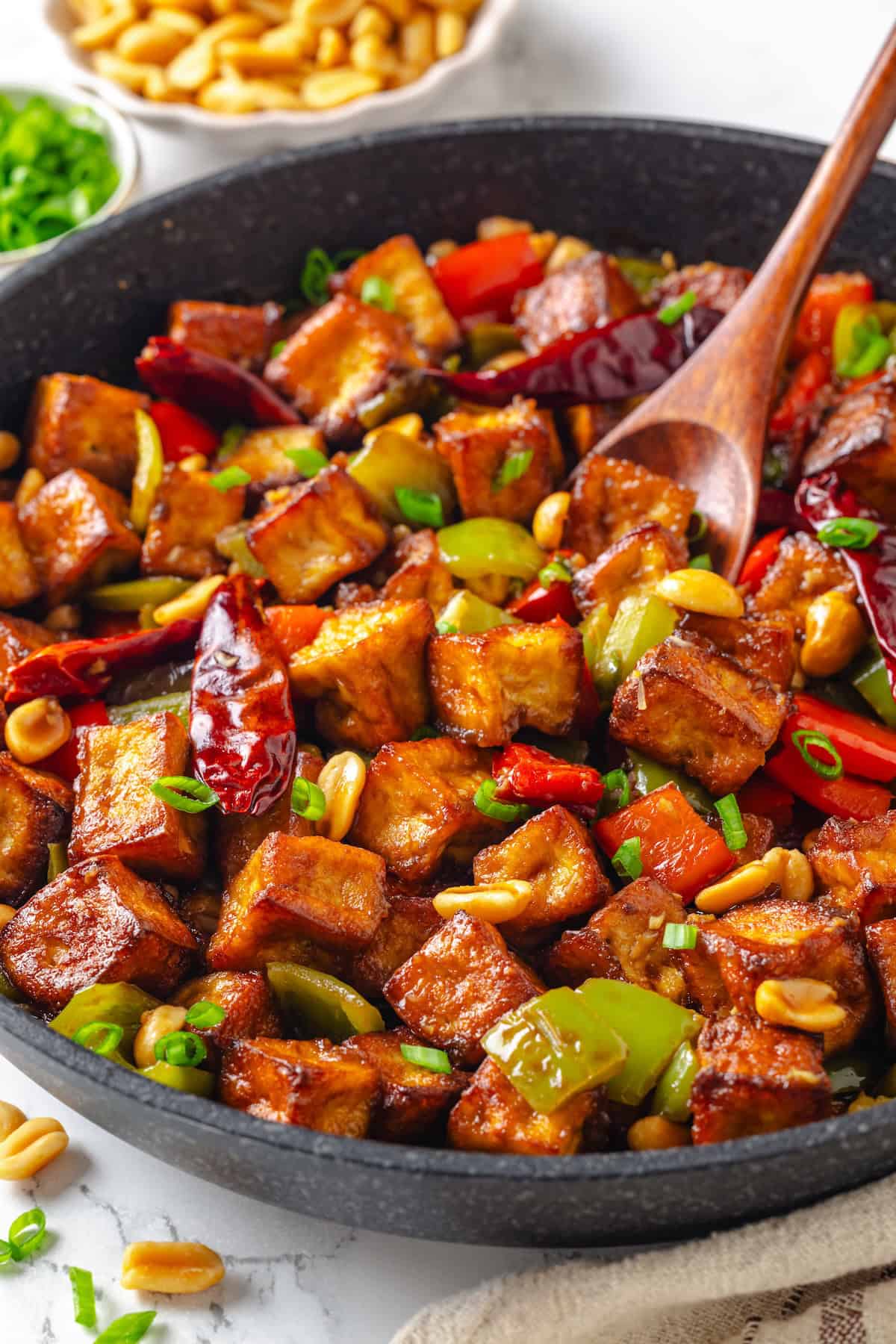 This crispy kung pao tofu is made with bell peppers, crunchy peanuts and a sweet-spicy sauce for a dinner that's even better than takeout! Wooden spoon stirring kung pao tofu in skillet.