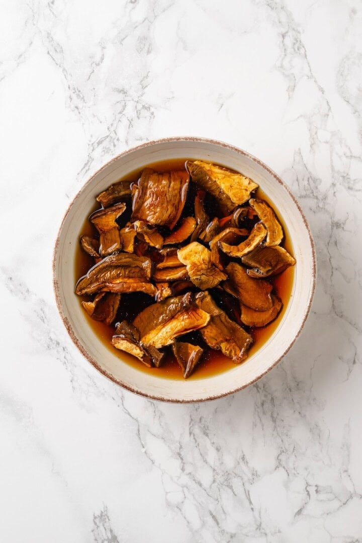 This homemade mushroom ravioli is filled with porcini, cremini and ricotta, then tossed in a creamy vegan Parmesan sauce. Restaurant-worthy! Porcini mushrooms in bowl with water.