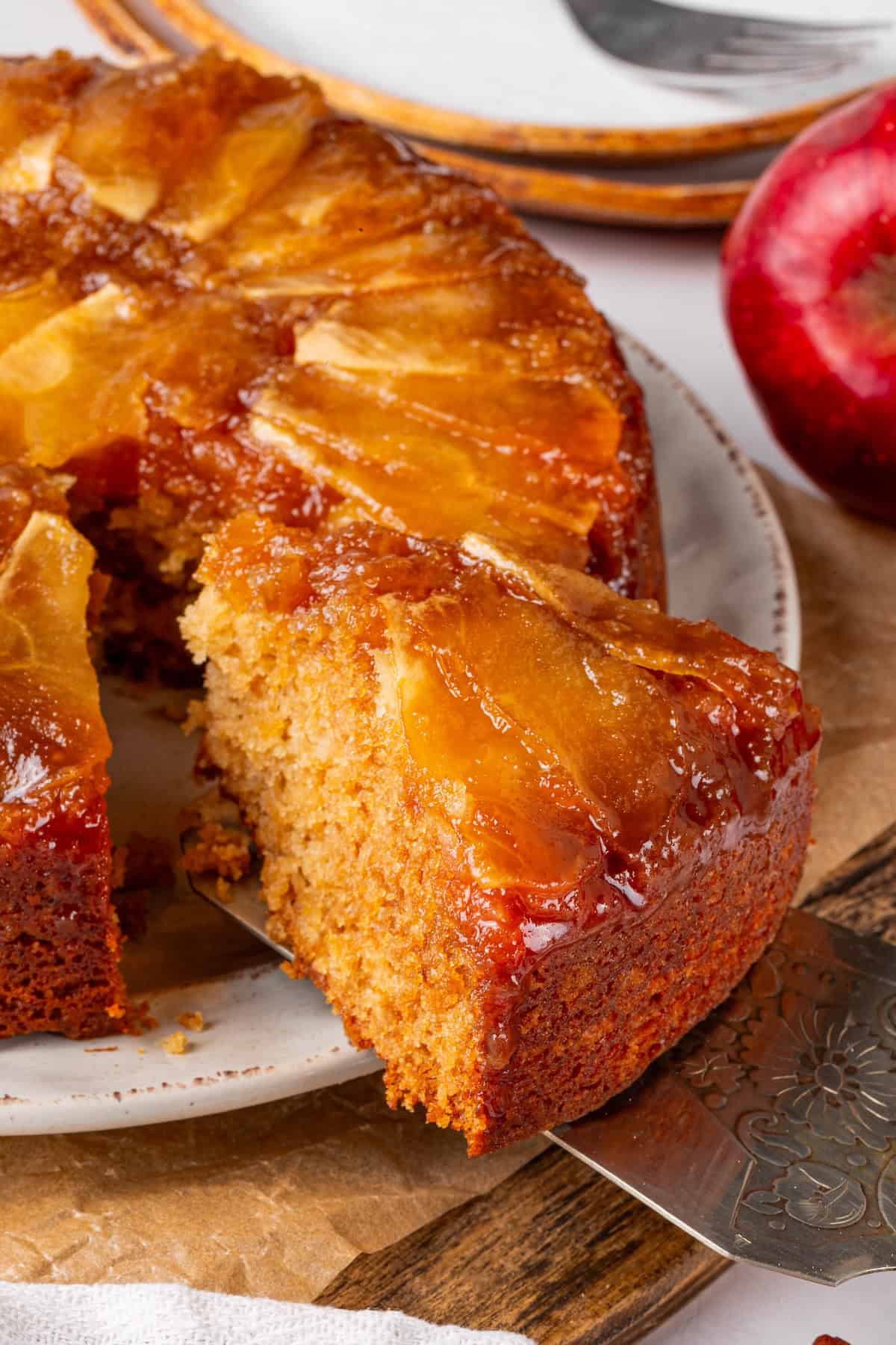 This easy upside down apple cake features tender, spiced cake topped with sticky caramel apples. It’s cozy, simple and perfect for fall! Removing slice of upside down apple cake from cake plate.