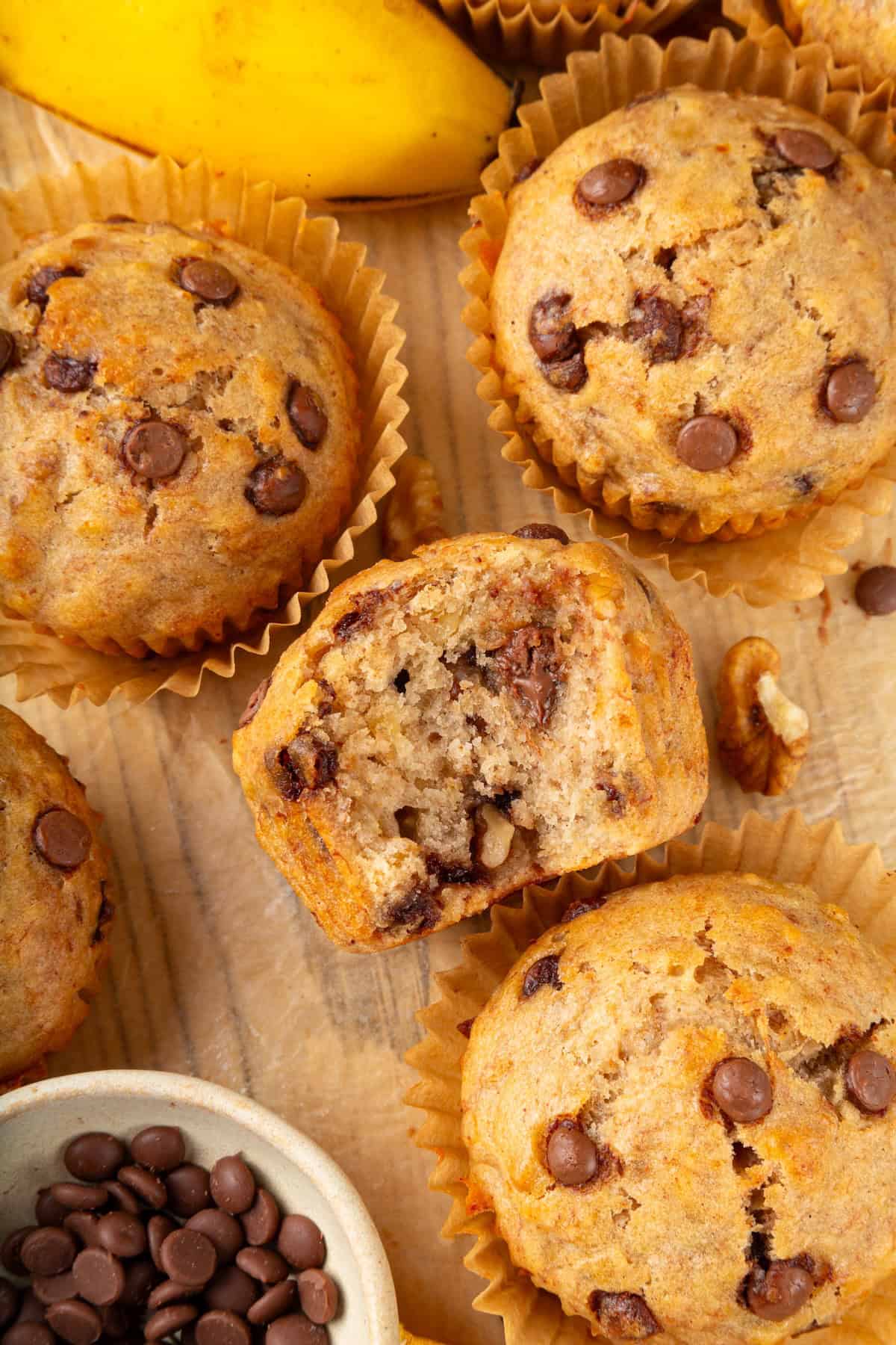 These vegan banana chocolate chip muffins bake up soft, moist and full of chocolate and crunchy walnuts. Perfect for breakfast and snacks! Vegan banana chocolate chip muffins on wooden board with one bitten into to show texture.